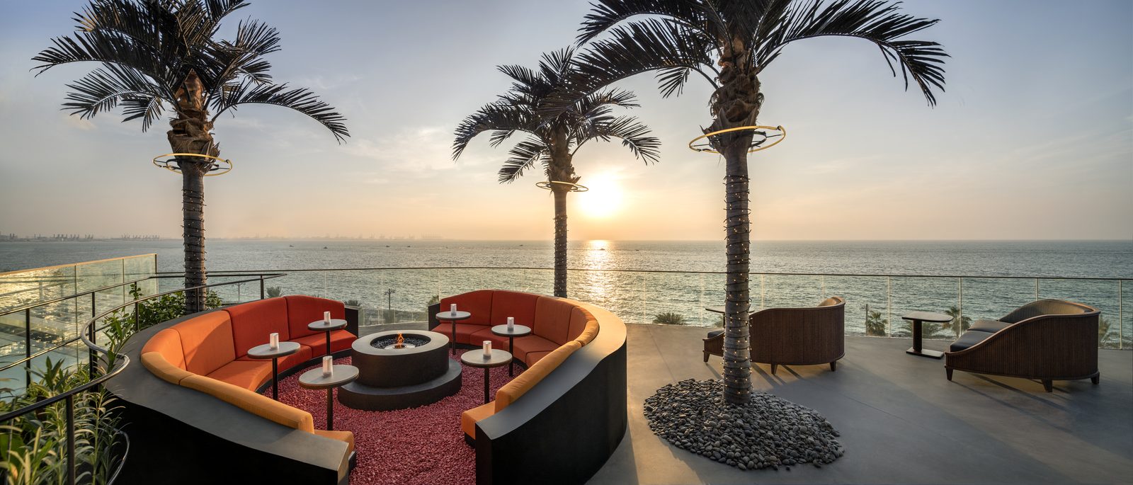 Curved orange sofas around a fire pit on the SoBe terrace at W Dubai - The Palm, with two palm trees and the sea at sunset