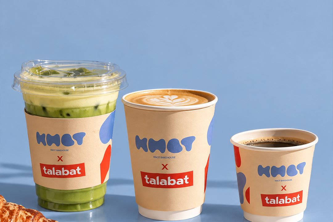 Three Knot Bakehouse and talabat coffee cups, a matcha drink, a latte and an espresso, on a blue background beside a croissant