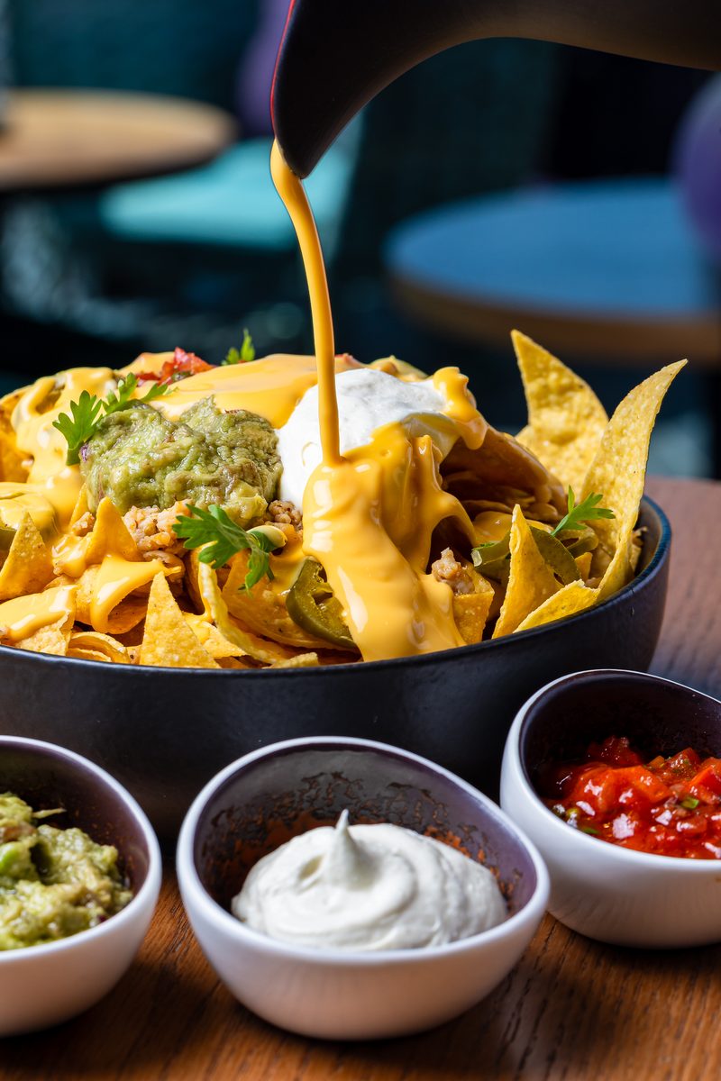 A bowl of nachos with cheese sauce being poured over guacamole, sour cream and salsa