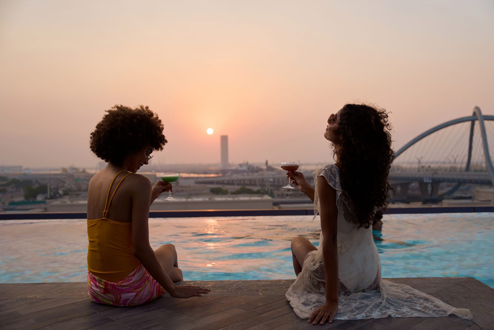 Two women sitting at the edge of a rooftop pool at sunset, with a bridge and the skyline behind