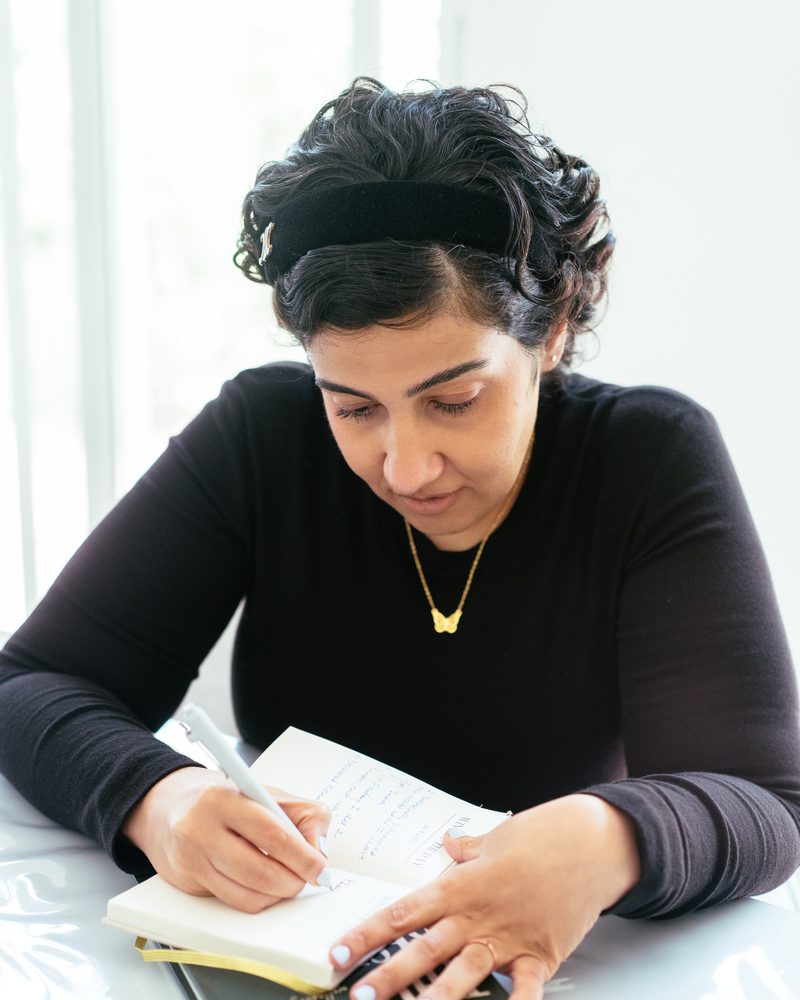 Rucha Daryani writing in a notebook at a white table, with a mug and a second book beside her