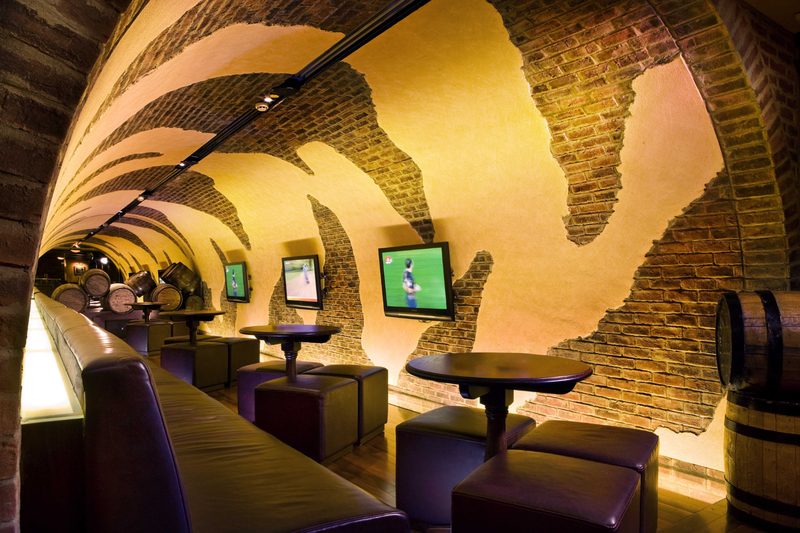 A curved brick-vaulted bar with leather seating, barrels and wall-mounted screens showing sport