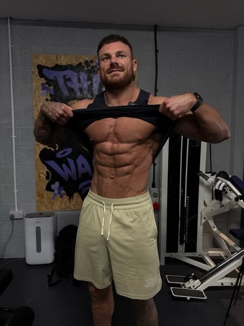A man with a beard and tattooed arms, in a black vest and sand-coloured shorts, pulling up his top in a gym with a graffiti-style poster and weight machines behind him