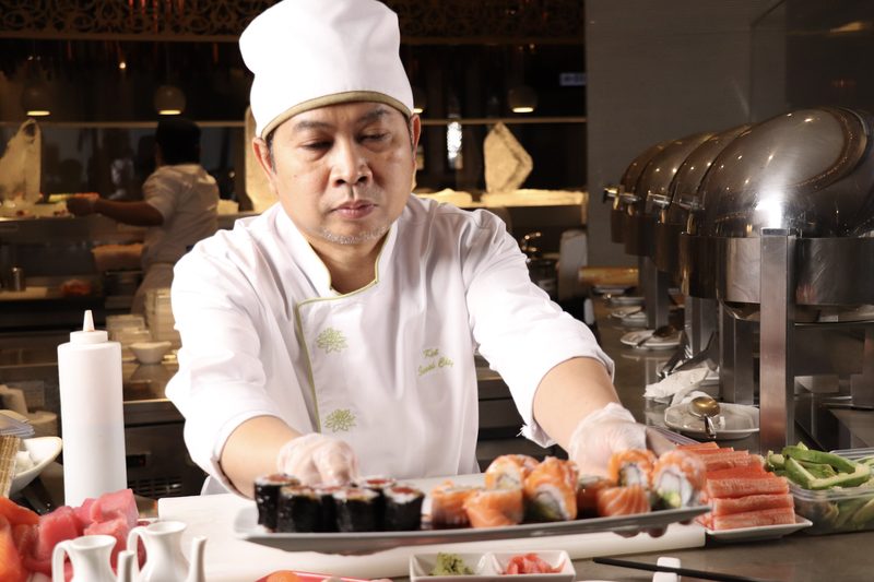 A chef in a white uniform at a sushi counter, squeezing sauce over a plate of sushi rolls with more rolls and fish on the counter