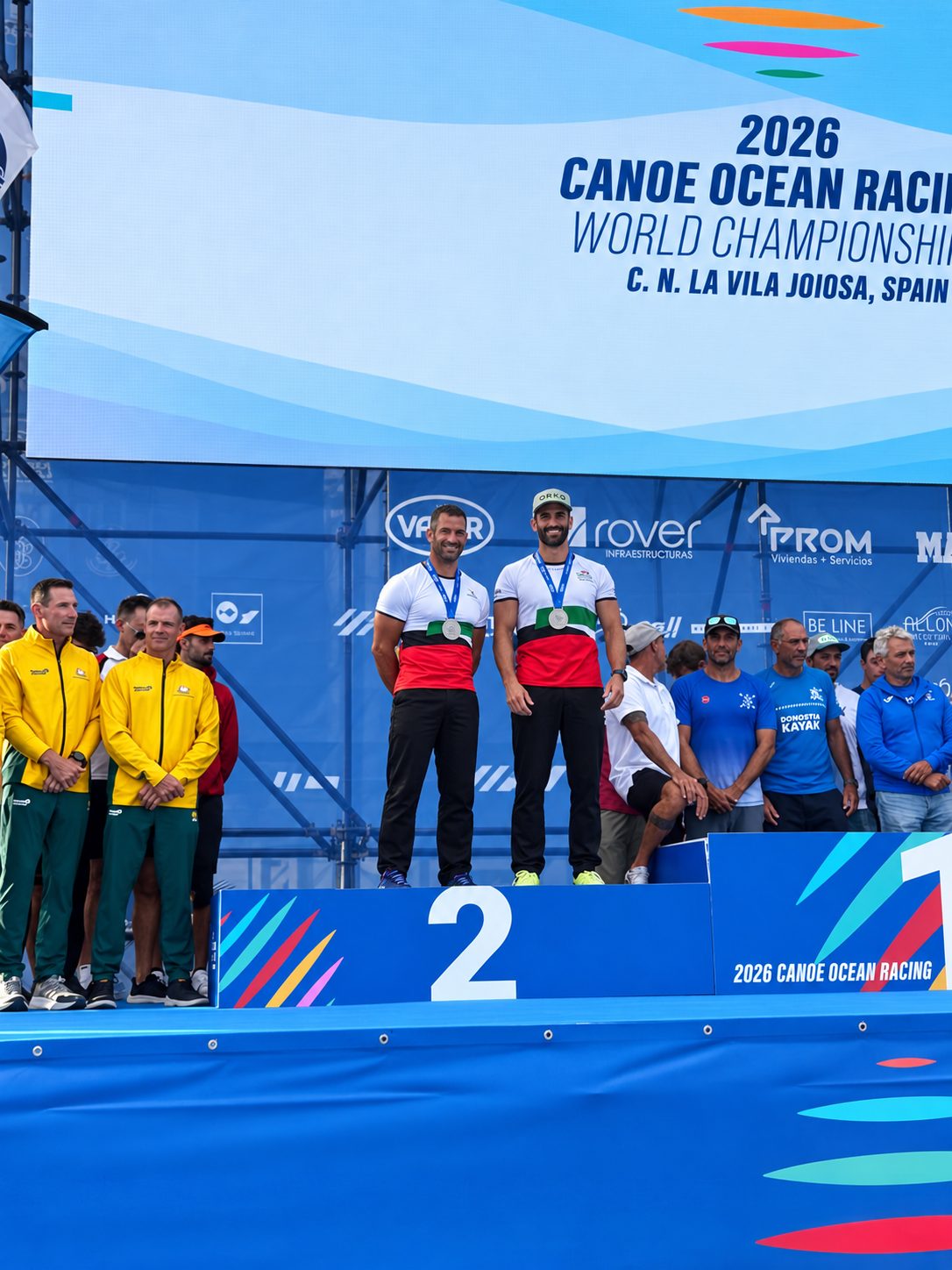 Two athletes in white and red kit stand on the second-place podium wearing silver medals, in front of a 2026 Canoe Ocean Racing World Championships banner in La Vila Joiosa, Spain
