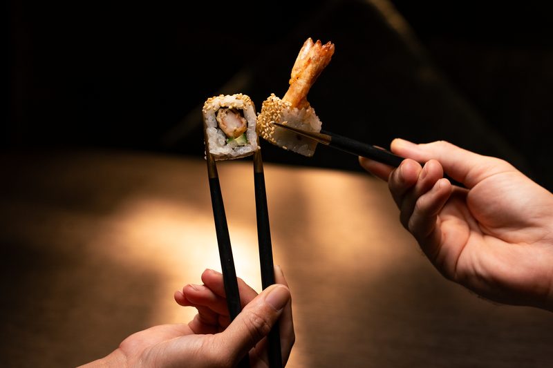 A hand holding chopsticks over a sushi roll topped with tempura prawn