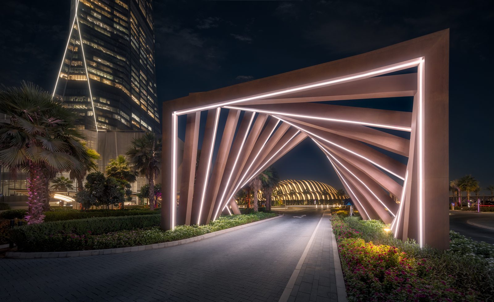 A lit geometric archway over a road at the entrance to Uptown Dubai at night, with a tower rising behind it