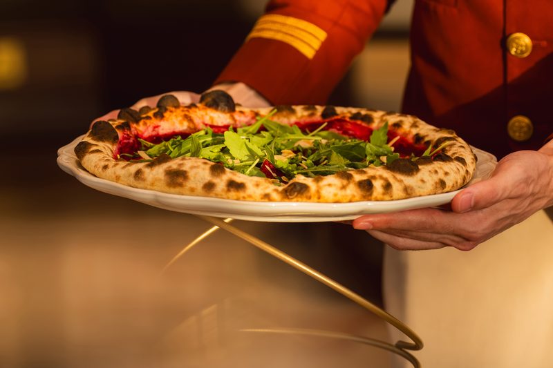 A server in a red jacket presenting a pizza topped with fresh rocket