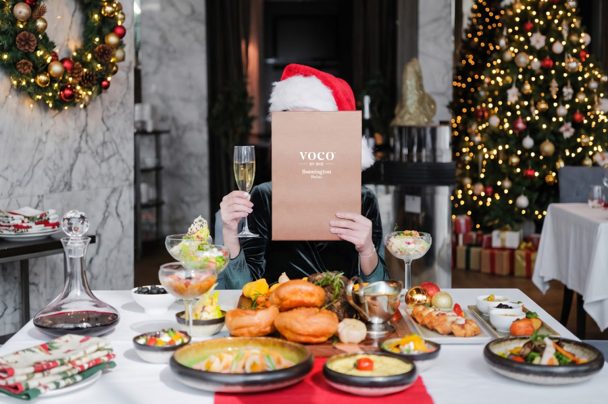 A festive table of dishes and a champagne flute, with a diner holding a menu in front of their face, beside Christmas trees and a wreath