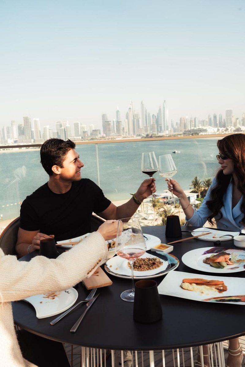Diners raising glasses at a terrace table with the Dubai skyline and the sea behind them, from the W Dubai The Palm release
